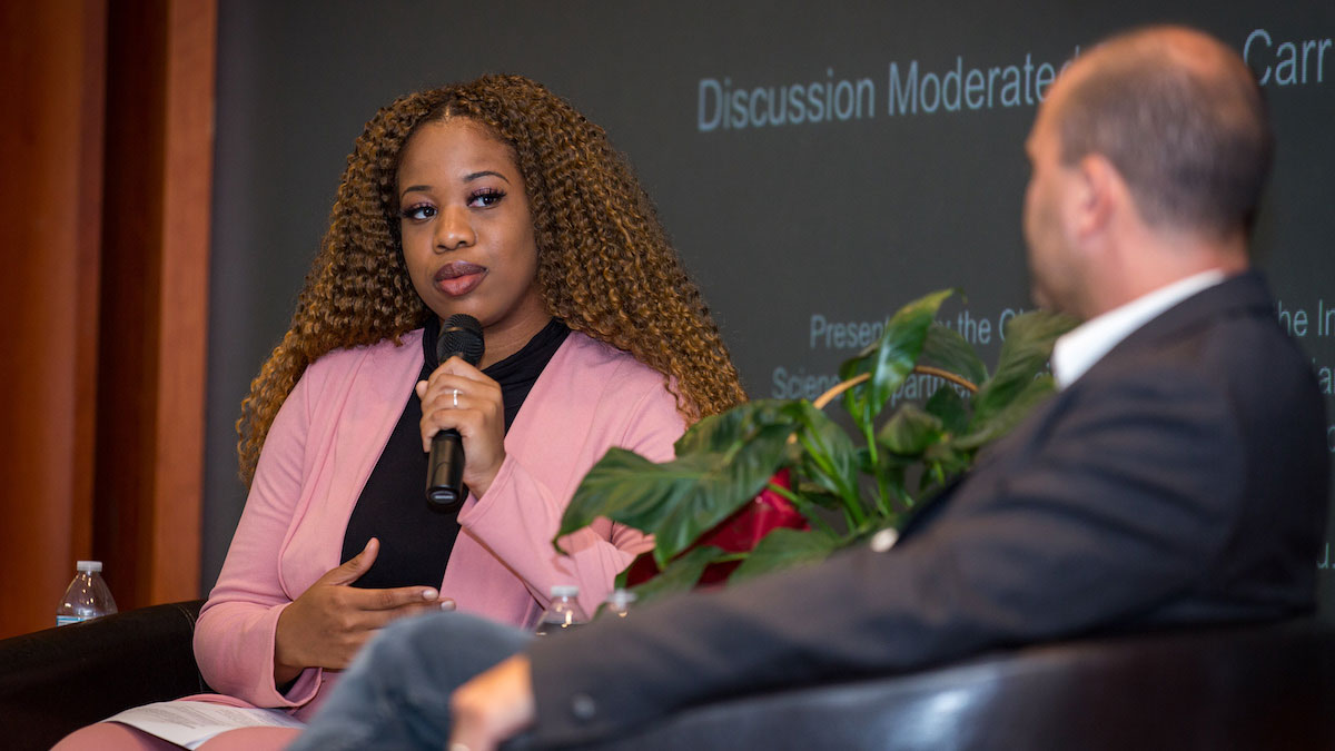 A student in professional attire holding a microphone and talking with a guest speaker in a lecture hall