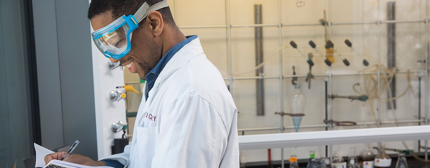 A student in a laboratory wearing a science robe and goggles and taking notes