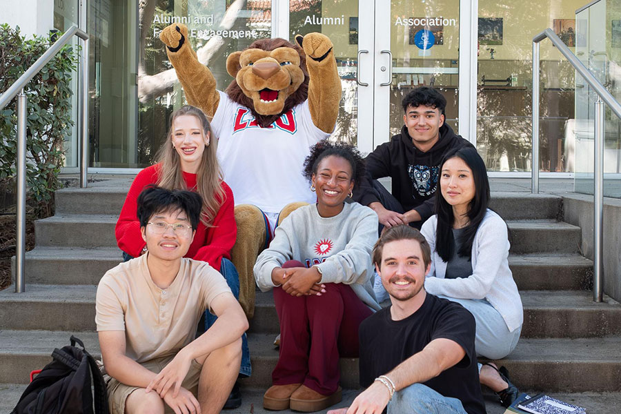 Students and Iggy the mascot smile on the steps in front of the Alumni Welcome Center on LMUs campus